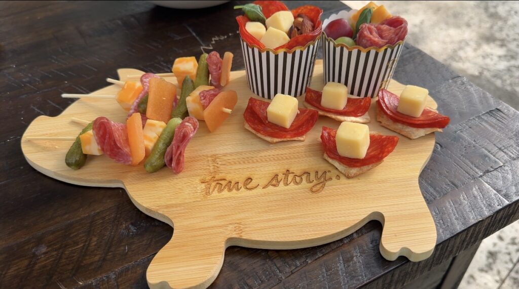 Charcuterie board on a guitar-shaped wooden board with skewers of cheese and vegetables, and pepperoni bites in striped cups.