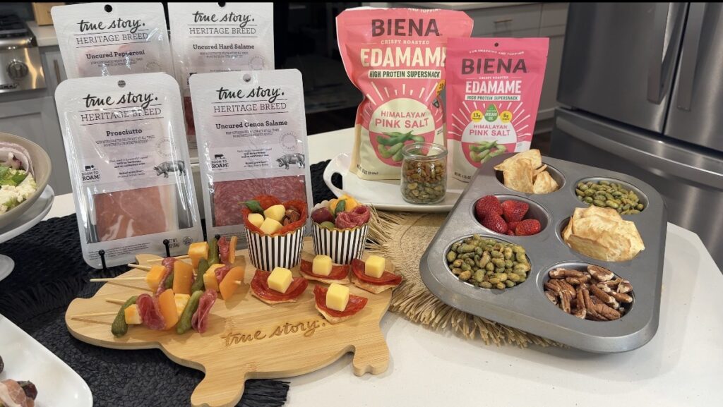 Snack spread: skewered cheese and meat bites on a wooden board and small cups with fruit, plus a muffin-tin of nuts and raspberries; true story Heritage Breed packs visible in the background.