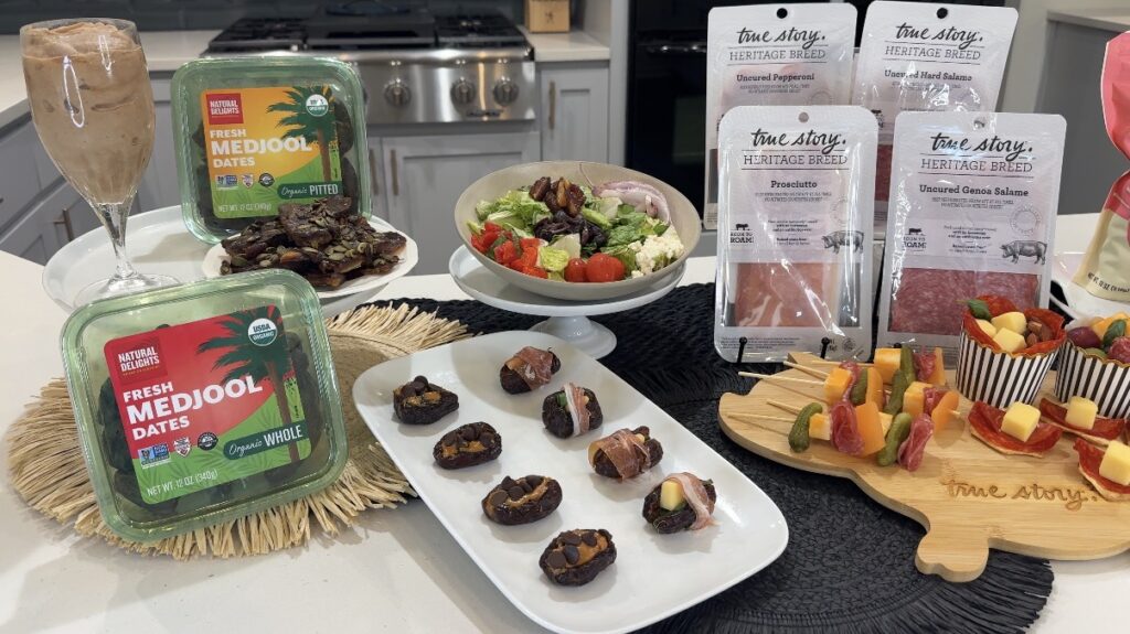 Assorted appetizers on a kitchen island: Medjool dates in green containers, bite-size date stuffed treats on a white plate, a bowl of leafy salad with tomatoes, and fruit-and-cheese skewers with packaged True Story Heritage Breed meats in the background, plus a tall glass of iced coffee on the left.